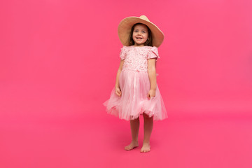 Portrait of cute little girl in straw hat and pink dress in the studio on pink background. Copy space for text.