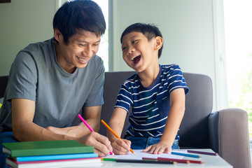 An Asian father is teaching and enjoys studying with his son, drawing crayons, sitting on the sofa....