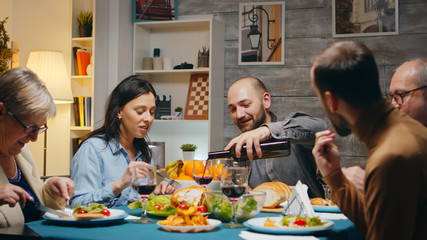 Husband pouring red wine to her beautiful wife at multigenerational family dinner.