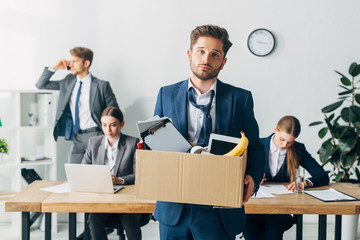 Fired man in suit holding cardboard box with papers and banana near colleagues working in office