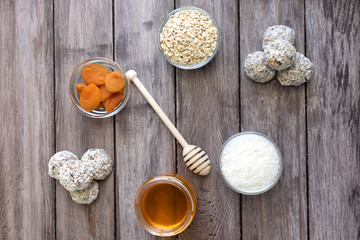 Ingredients for making protein balls: coconut, honey, oatmeal, dried apricot in small bowls and cooked energy balls in heaps on a wooden background