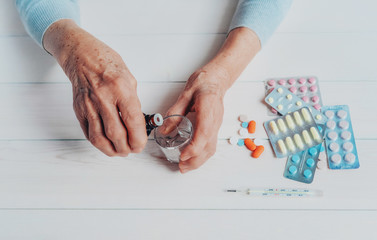 Senior hands with medicine bottle putting drops in a glass, pills, drugs, thermometer on a table. Woman wrinkled hands, colorful tablets, wooden background. Health care for the elderly, old age.