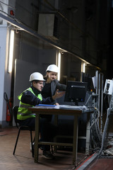 Plakat Portrait of a female factory manager in a white hard hat and business suit and factory engineer in work clothes. Controlling the work process in a factory.