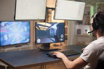 Back shot of man playing shooter game on computer with multiple monitors.