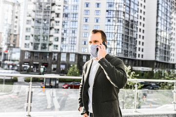 A guy in smart casual wear with a surgery protective mask on the face is talking on the phone in the downtown. Stay safety during quarantine, epidemia