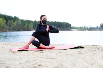 Fototapeta premium girl with dark hair and fair skin in black sportswear and a black protective mask goes in for sports on pink carimat on the sand by the lake in the forest