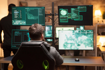Rear view of hacker in front of computer with multiple screens in dark room.