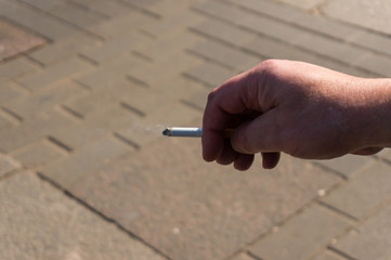 A burning cigarette in a male hand on the background of a city pavement. Bad habit.