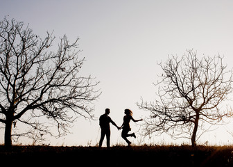 Silhouettes of a couple of man and woman outdoors, against sunset sky, walking between trees.