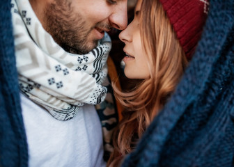 Close-up of a young couple of man and woman, wearing warm clothes, hugging on a winter day under a big knitted scarf..