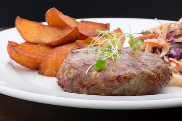 A burger with baked potatoes, cabbage and a salad in a ceramic plate on a wooden table