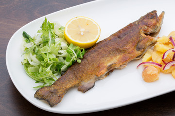 A roasted trout with potatoes, onions and lettuce  in a white ceramic plate on a wooden table