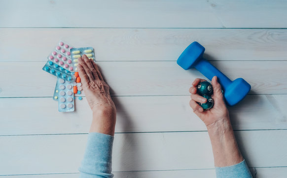 Senior wrinkled hand pushing back pills and thermometer, dumb-bell on a table. Another old woman hand with chinese health exercise baoding balls. Elderly health care, old age, healthy living concept.