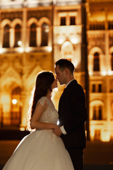 Fototapeta premium Bride and groom hugging in the old town street. Wedding couple walks in Budapest near Parliament House in night. Happy romantic young couple celebrating their marriage. Wedding and love concept.