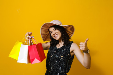 Portrait of a cheerful young Asian woman with  hat holding shopping bags and presents gestures isolated over yellow background