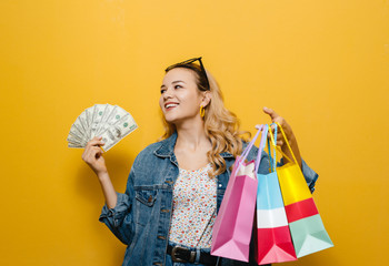 Image of a excited  young blonde girl holding banknotes and shopping bag  over yellow background