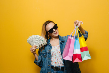 Image of a excited  young blonde girl holding banknotes and shopping bag  over yellow background