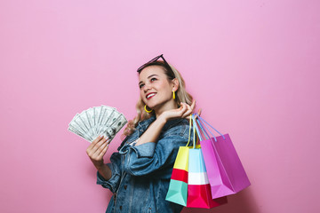 
Image of a happy blonde girl thinking about something and holding banknotes and shopping bags on pink background