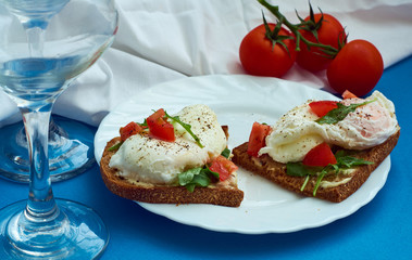 Poached eggs on sandwich of rye bread with sauce, tomatoes and arugula in white plate on blue background.