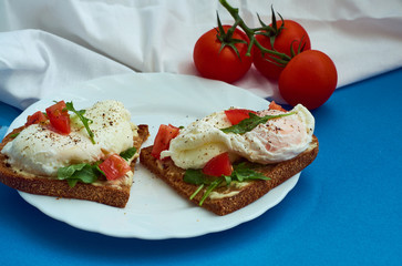 Fototapeta na wymiar Poached eggs on sandwich of rye bread with sauce, tomatoes and arugula in white plate on blue background.