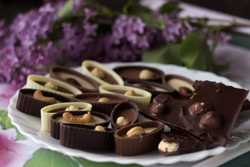 Bouquet of lilac flowers and chocolate candies with filling and nuts, chocolates with nuts on a plate, background. Sweets made of black, milk and white chocolate with hazelnuts, almonds and cashews