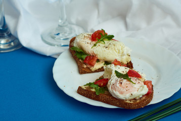 Poached eggs on sandwich of rye bread with sauce, tomatoes and arugula in white plate on blue background.