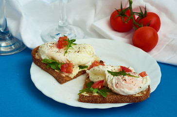 Poached eggs on sandwich of rye bread with sauce, tomatoes and arugula in white plate on blue background.