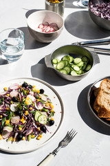 Photo of a fresh spring salad on a white porcelain plate and vegetable mix of ingredients in metal bowl on a white rustic background sunlight with shadows
