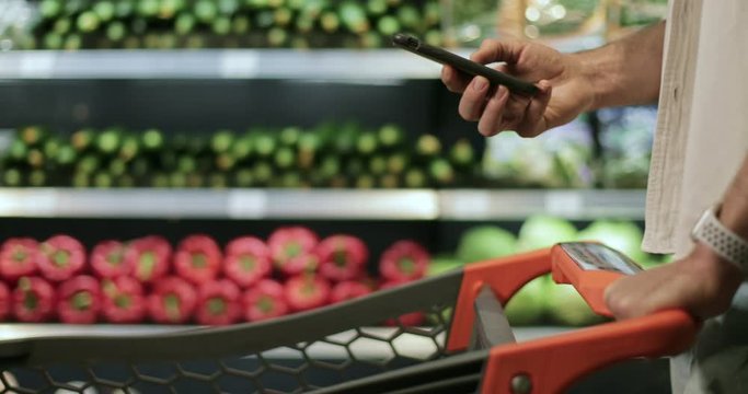 Crop view of man hand holding smartphone and srolling screen in supermarket. Guy using smartphone while walking and pushing shopping cart near vegetables shelves. Blurred background