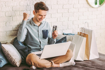 Happy male shopper shopping online with laptop, hold credit card in hand and smiling on couch....