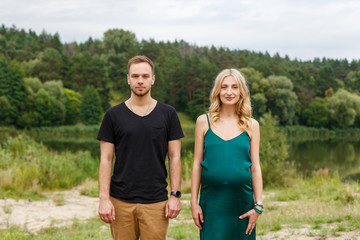 Pregnant woman in green dress and her husband in black t-shirt standing near each other and looking to the camera. Stylish interesting couple on the background of nature. Young family and pregnancy