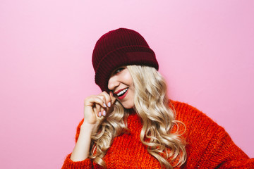 Portrait of a young blonde girl wearing a cap and is dressed in a red sweater and showing fashion moves over pink background