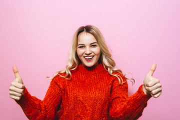 Portrait of a blonde girl who is dressed in a red sweater and smiles in the room and gestures on a pink background