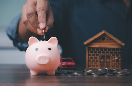 Man hand putting money coin into piggy for saving money wealth and financial concept, with wooden home and red car in back.