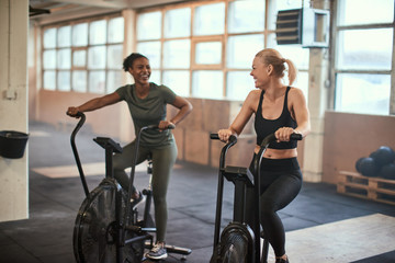 Fototapeta premium Two laughing young woman riding stationary bikes in a gym