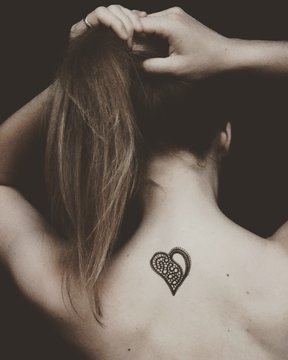 Rear View Of Topless Woman With Heart Shape Tattoo On Back Against Black Background