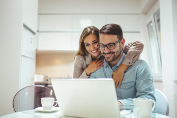 Pleasant family couple sitting at big wooden table in modern kitchen, looking at laptop screen. Happy young mixed race married spouse web surfing, making purchases online or booking flight tickets.