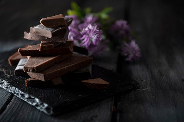 Pyramid of broken chocolate pieces on black background next to violet flower.