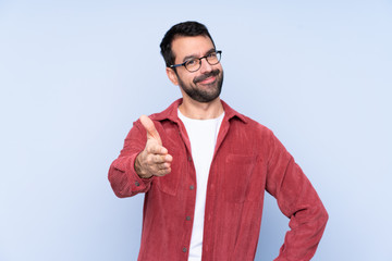 Young caucasian man wearing corduroy jacket over blue background shaking hands for closing a good deal