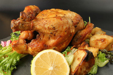 Whole roasted grilled chicken poultry bird with baked potato vegetable salad tomato lemon on black slate stone cutting board black background