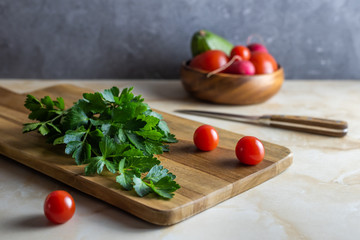 A fresh celery leaves lie on a wooden cutting board with a knife, on a marble kitchen table. Behind a bowl with vegetables, tomatoes, radish. With a gray concrete background.