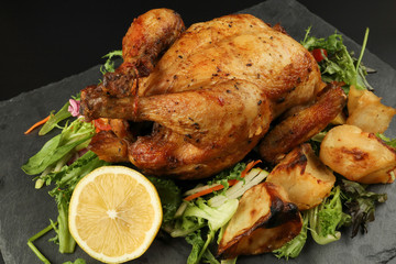 Whole roasted grilled chicken poultry bird with baked potato vegetable salad tomato lemon on black slate stone cutting board black background