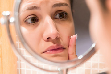 Young girl in front of a bathroom mirror putting cream on a red pimple. Beauty skincare and...