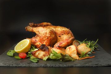 Whole roasted grilled chicken poultry bird with baked potato vegetable salad tomato lemon on black slate stone cutting board black background