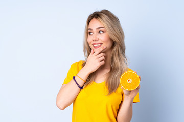 Teenager Russian girl holding an orange isolated on blue background thinking an idea and looking side