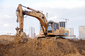 Obraz premium Yellow excavator during earthworks at construction site. Backhoe digging the ground for the foundation and for laying sewer pipes district heating. Earth-moving heavy equipment