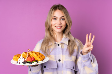 Teenager Russian girl holding waffles isolated on purple background happy and counting three with fingers