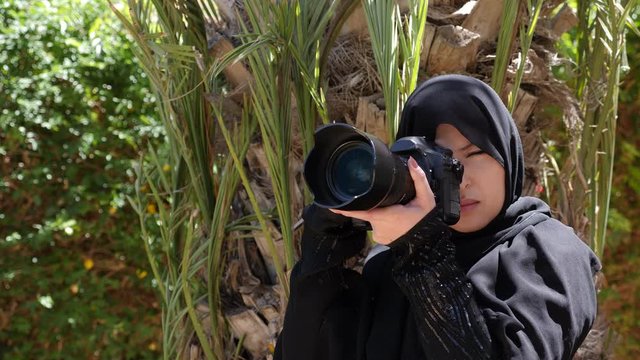 A beautiful Moroccan Arab Muslim woman with a DSLR camera filming photographing in a garden - turning the camera towards the viewer. Real-time footage.