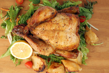 Whole roasted grilled chicken poultry bird with baked potato vegetable salad tomato lemon on wooden cutting board background