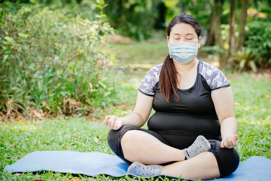 Asian fat girl doing yoga meditation at outdoor public green park and wear face mask for virus spreading protection.